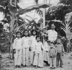 Lunar New Year (Tết) 1964 in front of the house in Quới Sơn. Má holds Lượng (6 months). Siblings from left to right: Huệ (Chị Tư), Lan (Chị Ba), Thanh (Chị Hai), Thơm (Chị Năm), Thượng (Anh Bảy), and Vân (Anh Sáu).